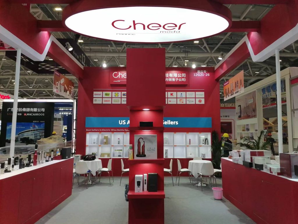 A red and white exhibition booth displays wine accessories, with tables, chairs, and an illuminated overhead sign.