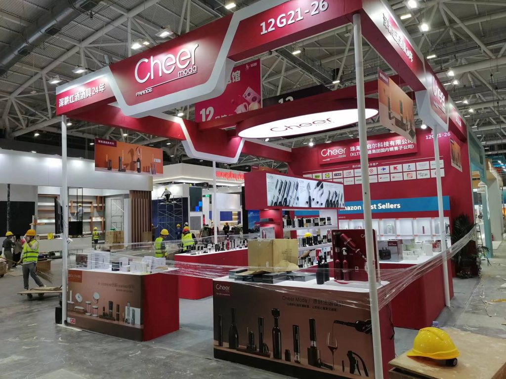 A red and white exhibition booth being set up, with displays of wine accessories, protective plastic, and workers in hard hats.