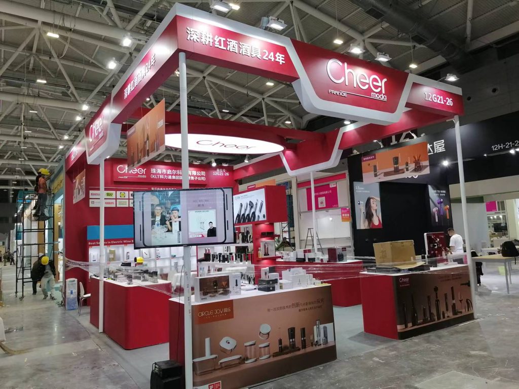 A red and white exhibition booth with screens and product displays is under construction, with workers and materials around it.
