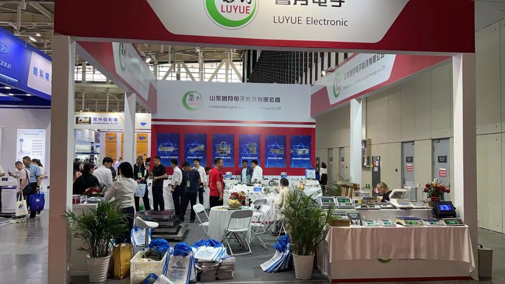 A busy trade show booth with many attendees and staff. Display tables show various scales, surrounded by plants and red-and-white branding.