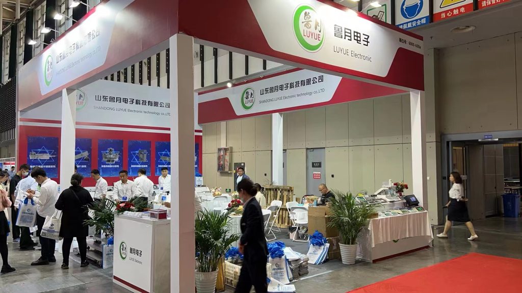 A trade show booth with red-and-white branding and product displays. A few people are present, including a woman walking by in the foreground.