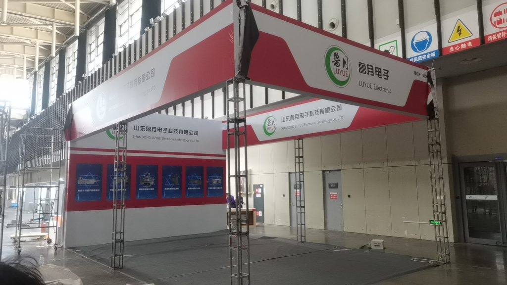 An empty trade show booth structure with red and white panels and visible metal trusses. The booth appears to be under construction or disassembly.