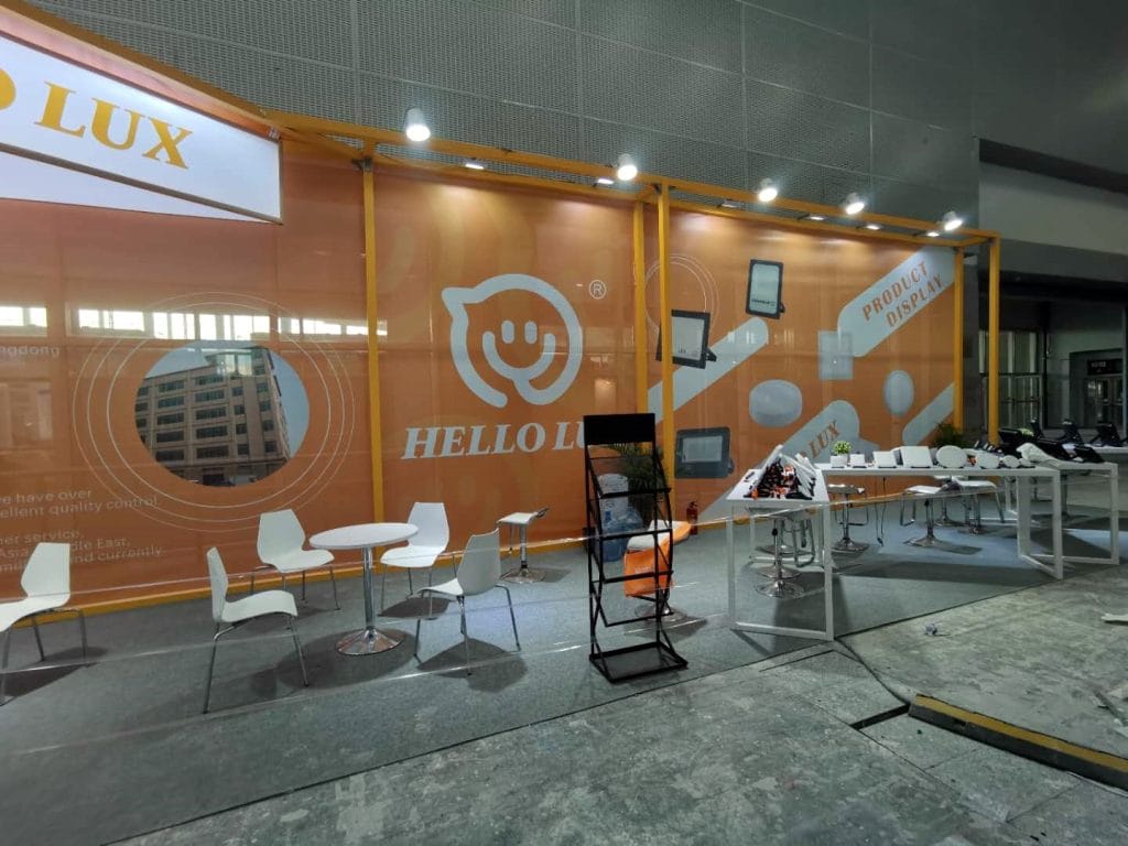 An orange and white exhibition booth during setup, with seating, product displays, and a magazine rack. Concrete floor visible.