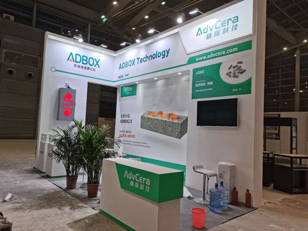 rade show booth being set up, featuring white and green branding. Displays show ceramic components, with potted plants and a counter in the foreground.