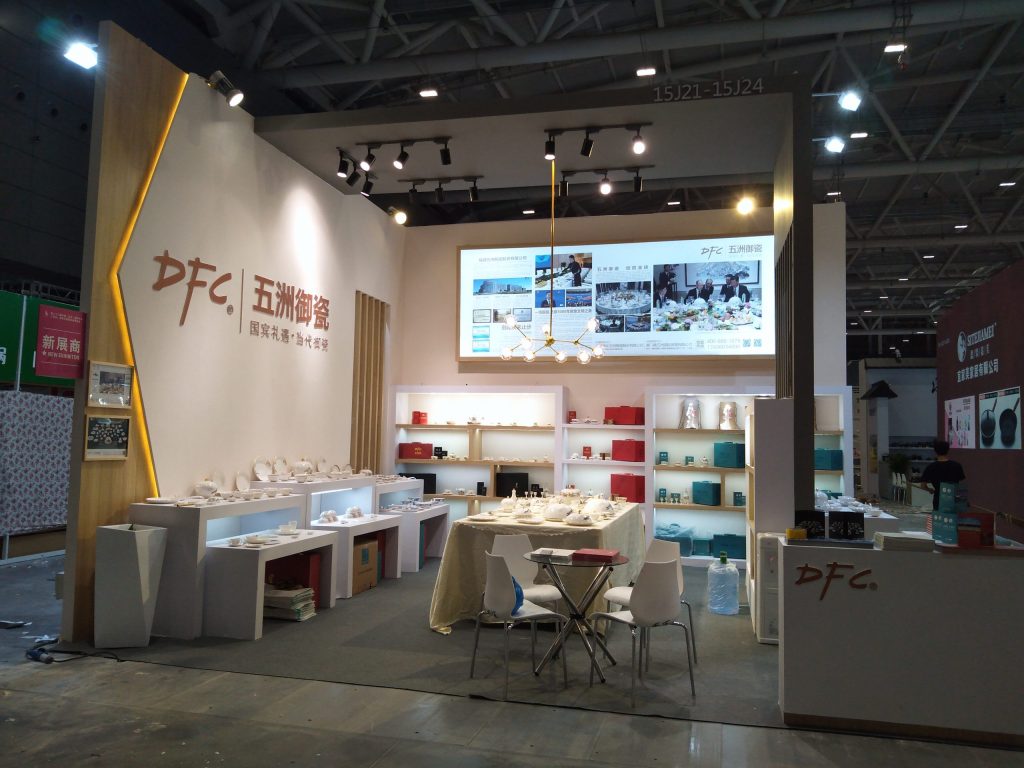 Modern exhibition booth with white shelves displaying porcelain and red items, a central table with chairs, a screen, and a contemporary chandelier.