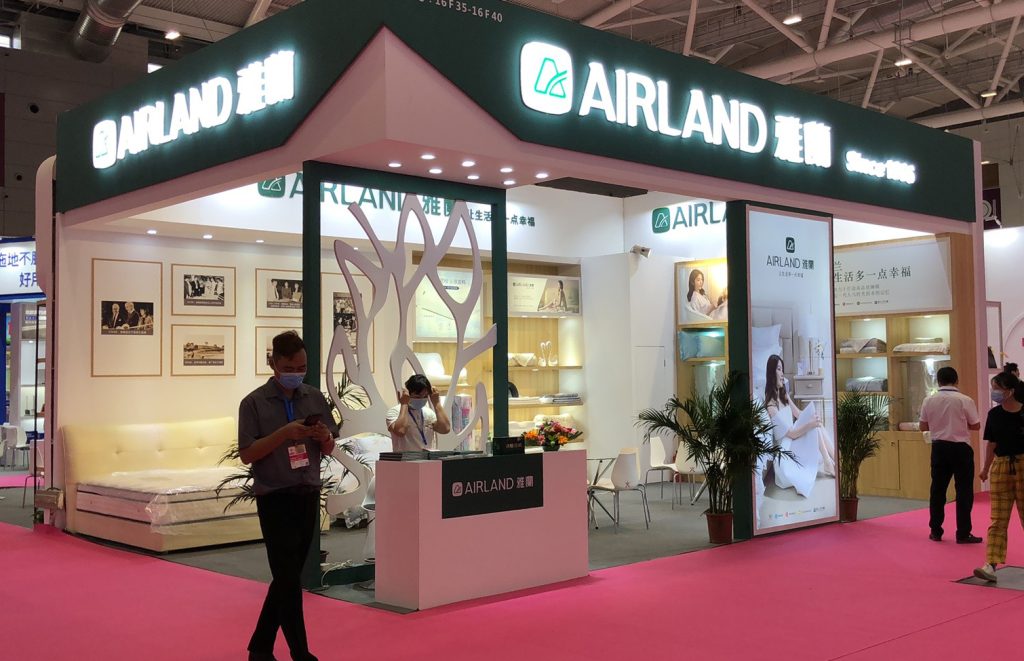Trade show booth with green trim, illuminated signs, and a pink floor. Displays feature mattresses and bedding, with people walking by.