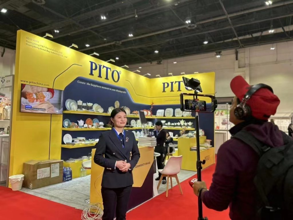 A woman in a dark suit speaks while being filmed by a person holding a smartphone on a stand at a busy exhibition booth.