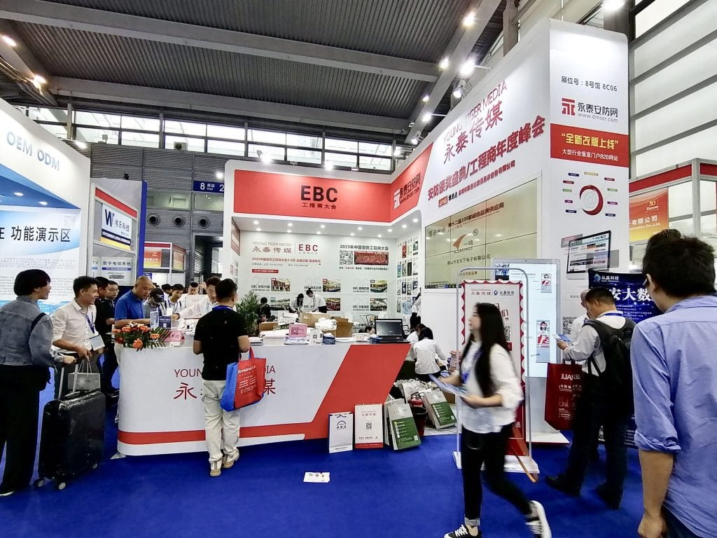 Lively trade show booth with many attendees engaging with displays and staff. An L-shaped counter and large display panels feature prominently.