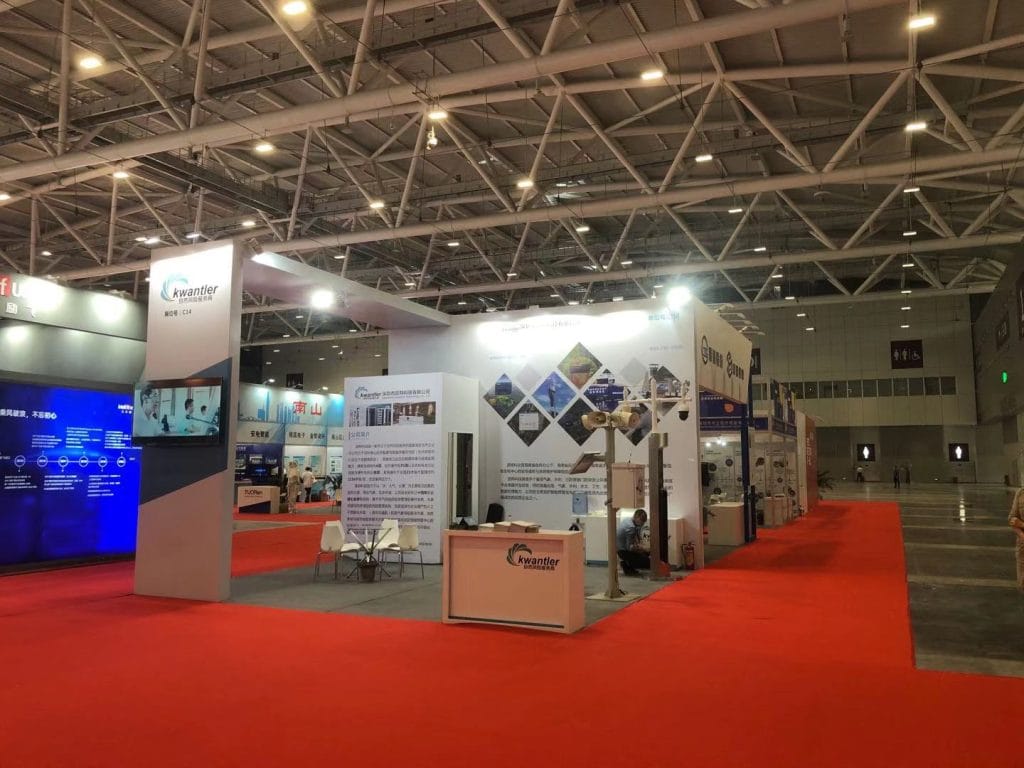 Wide view of a brightly lit trade show hall with multiple booths, a red carpet, and several people.
