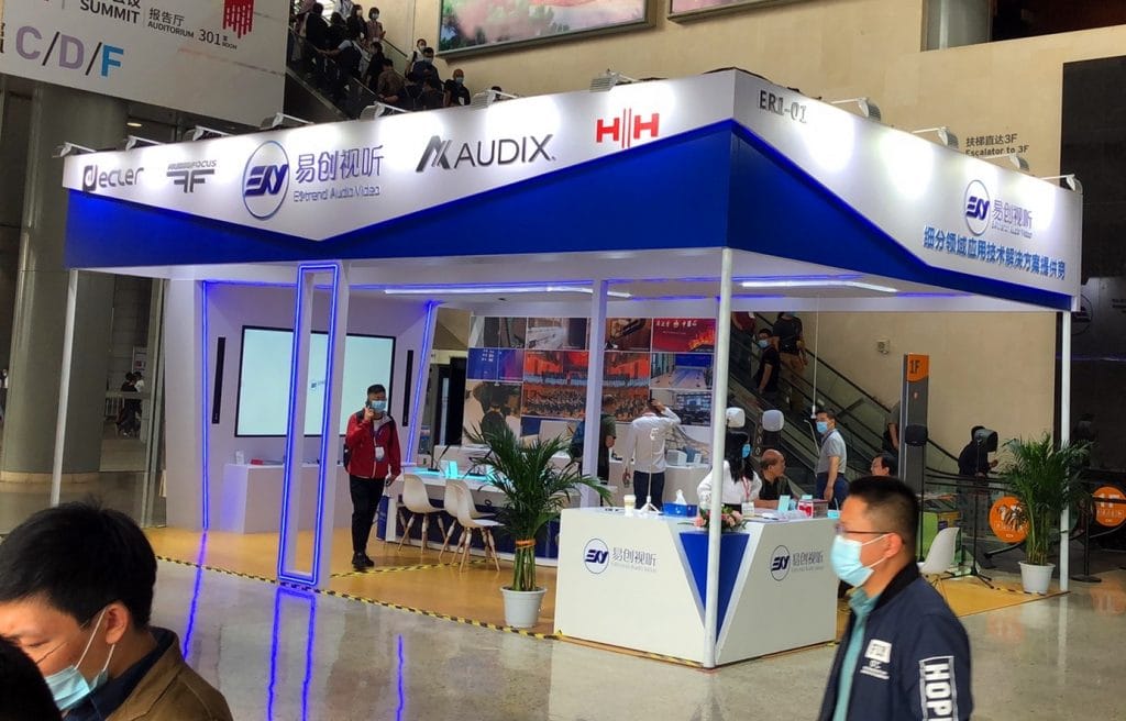 Blue and white exhibition booth with people walking around, some wearing masks, in a busy convention hall.