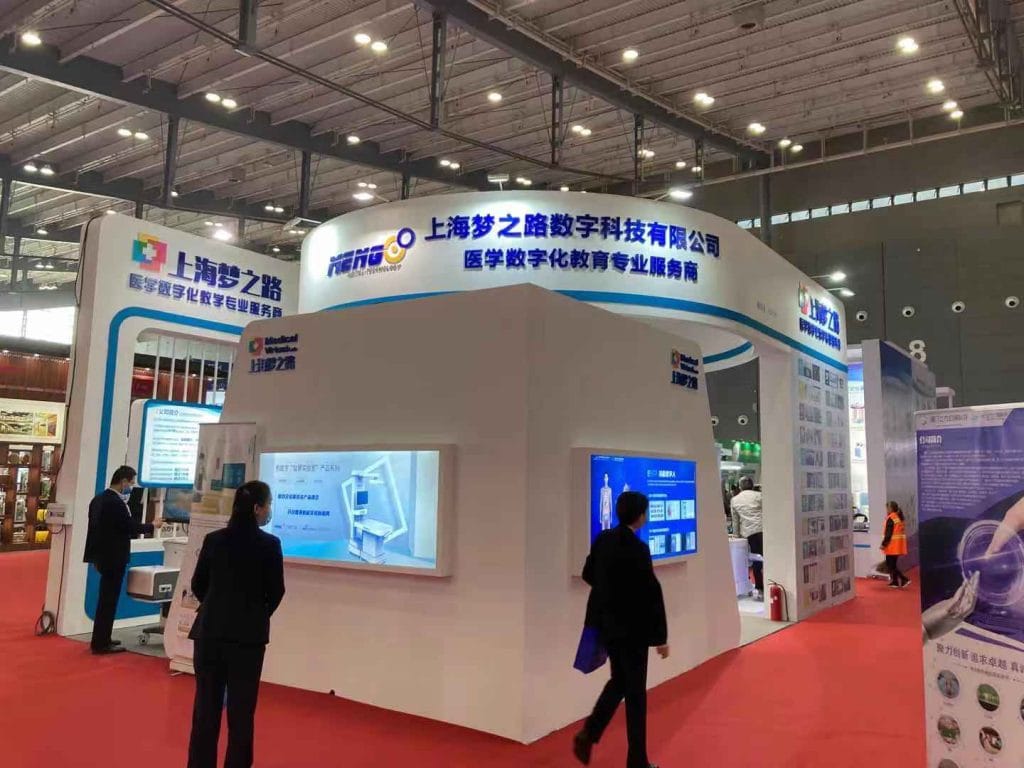 People walk past a large white and blue exhibition booth at a busy trade show with red carpet flooring.