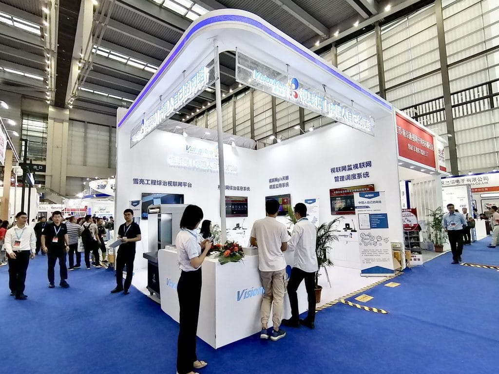 A bustling exhibition hall with a prominent white and blue booth where visitors interact with displays and staff. Other booths are visible.