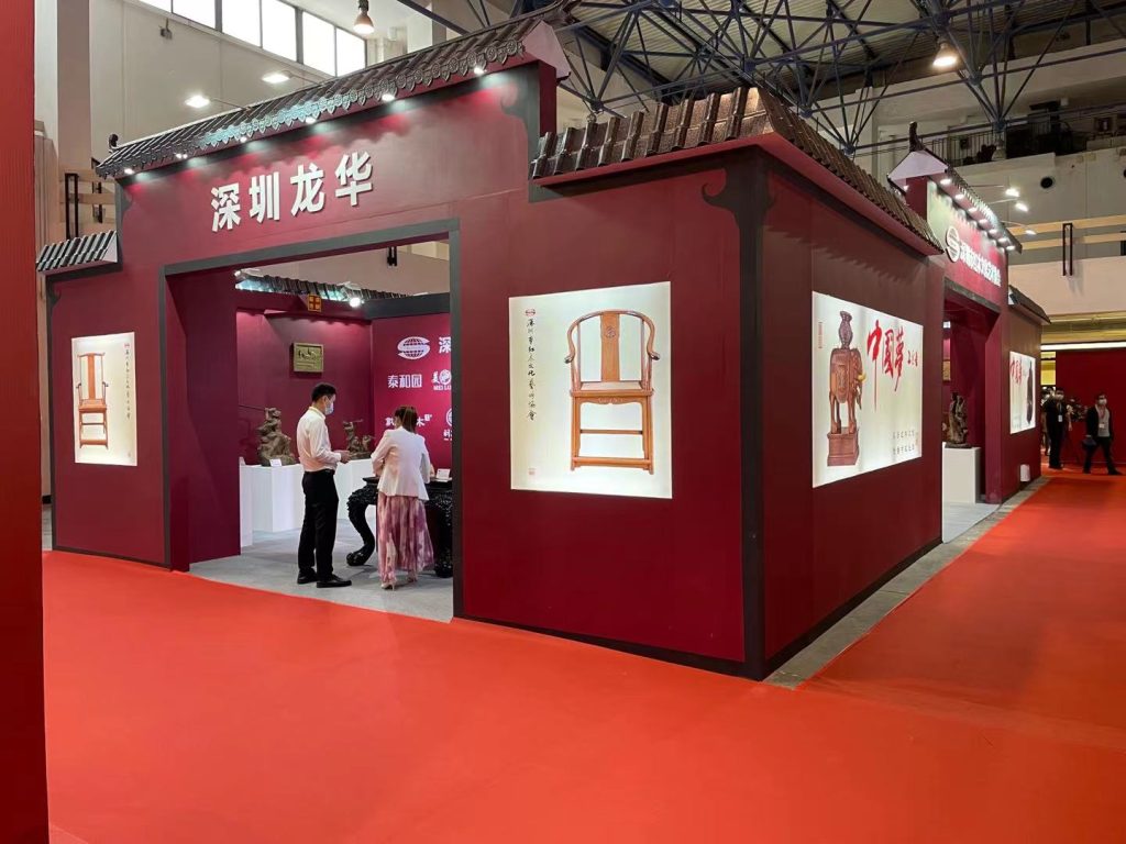 Red exhibition booth entrance with traditional Chinese roof. Display cases show a wooden chair and a statue. Two visitors are inside, facing exhibits on a red carpet.