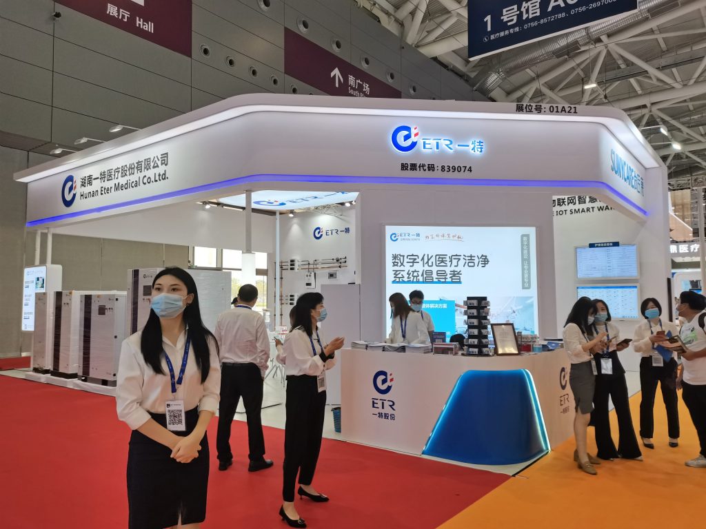 An exhibition booth with a red carpet, a reception desk, display screens, and multiple staff members, some wearing face masks.