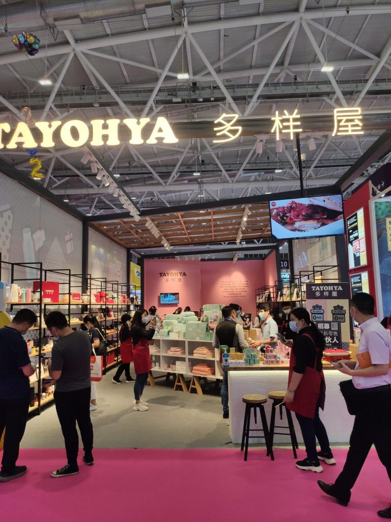 Crowded exhibition booth with a pink floor. Numerous masked visitors browse products on shelves and counters, assisted by staff in red aprons.