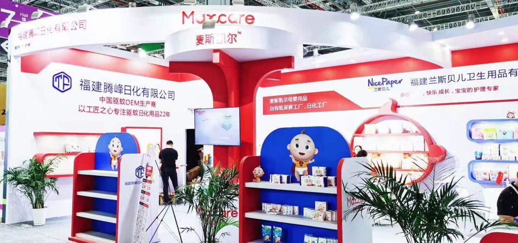 A vibrant red and white exhibition booth featuring display shelves, packaged products, and cheerful cartoon baby figures. Green plants decorate the space.