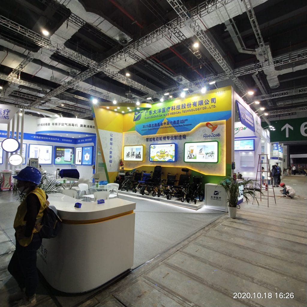 A brightly lit exhibition booth showcasing medical equipment, primarily wheelchairs, with large screens displaying products. People mill around in a large hall.