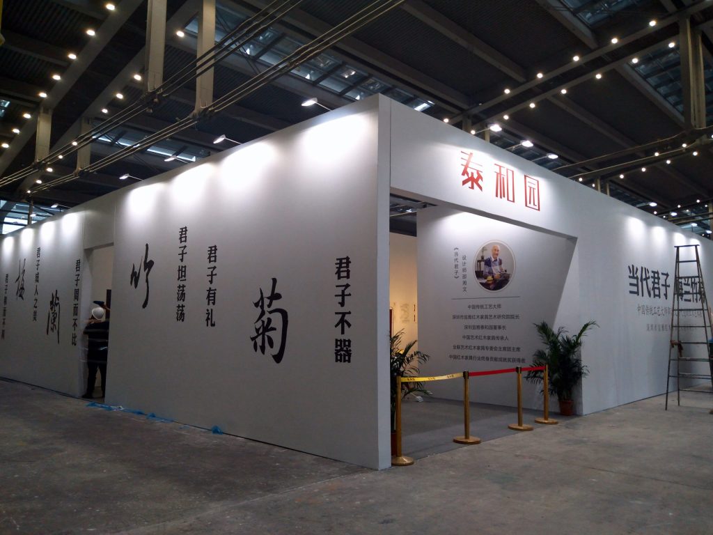 White exhibition booth with arched entrance, red and black Chinese characters, a portrait, and potted plants.