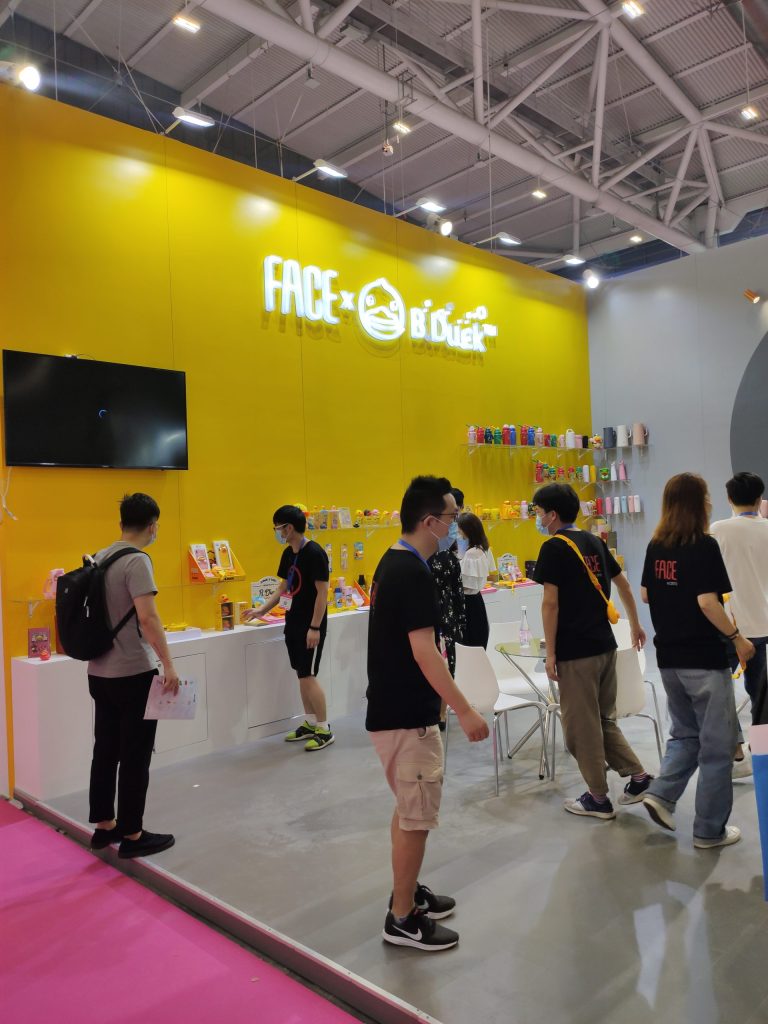 People wearing masks visit a vibrant yellow trade show booth, browsing various products displayed on a white counter.
