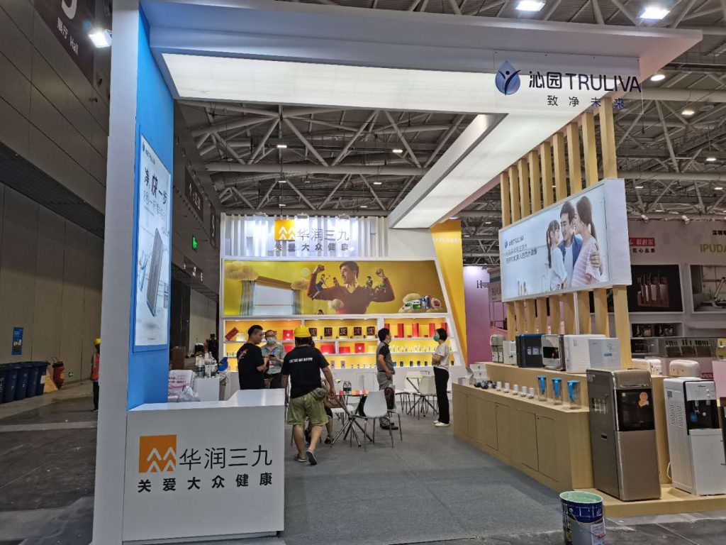 An exhibition booth with people gathered around tables and product displays. Some construction materials are visible on the left.