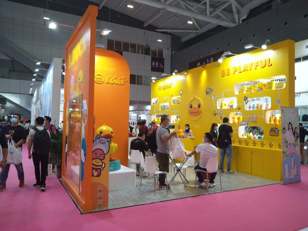 People gather at a vibrant orange and yellow trade show booth featuring cartoon duck characters and products.