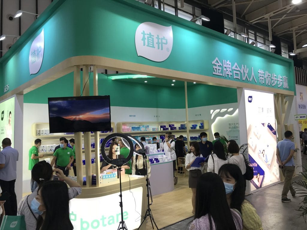 Crowded trade show booth with teal accents and white displays. Many masked people observe products; one person is filmed with a ring light.