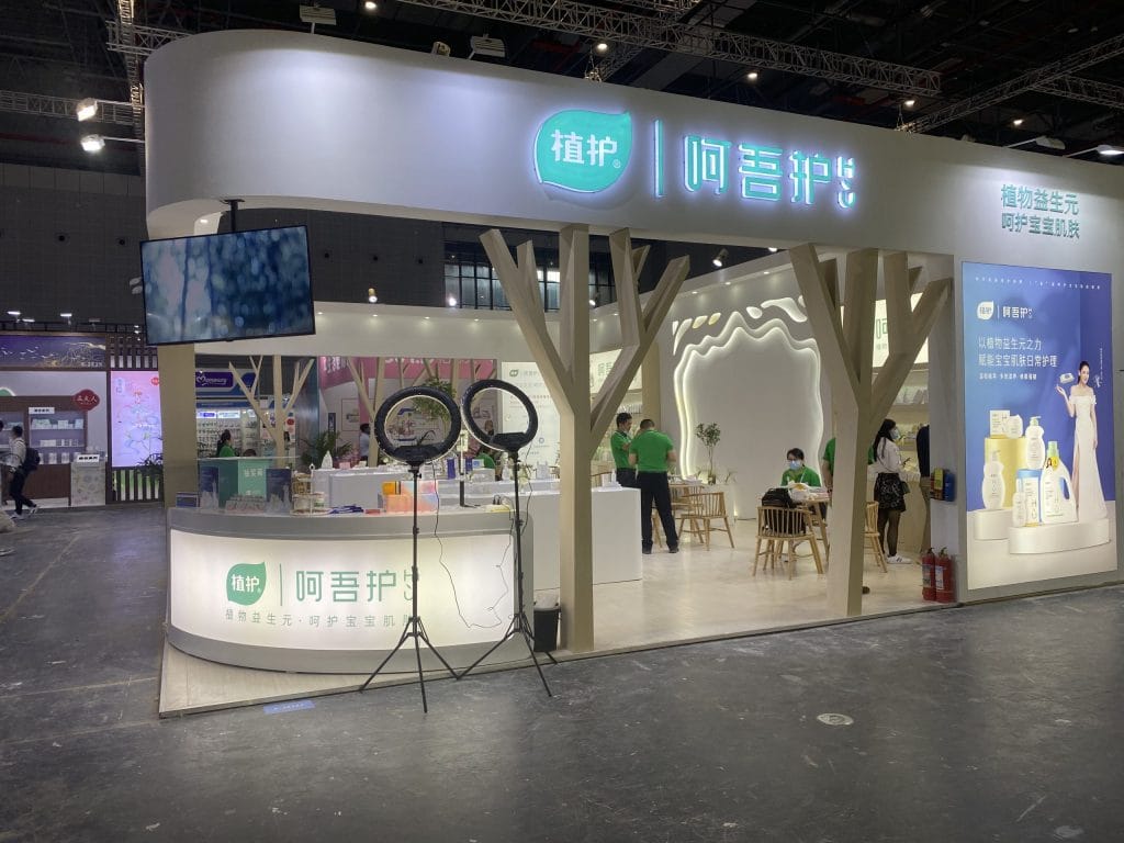 Live trade show booth: curved walls, glowing signs, tree-like pillars, and people. Ring lights visible.