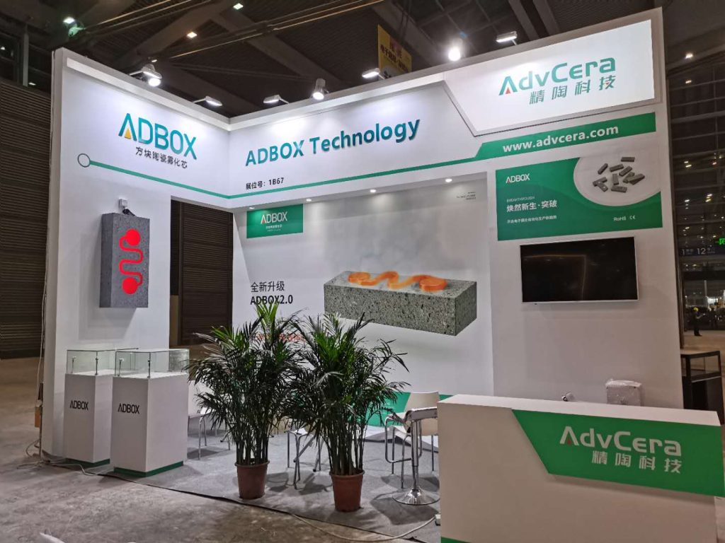 A white and green trade show booth, likely still under construction. It includes product displays, potted plants, chairs, and a front counter.