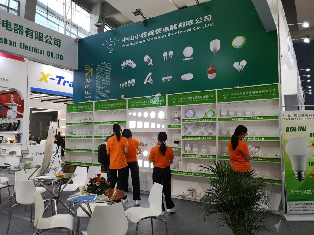 A busy exhibition booth with a green wall displaying various light bulbs and lighting products. Four women in orange shirts browse the displays.