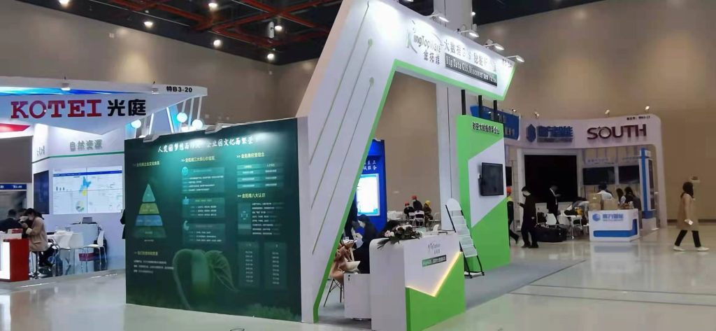 Real-life trade show floor with a white and green booth. People walk by, and a woman stands at the booth's reception desk. Info panels are displayed.