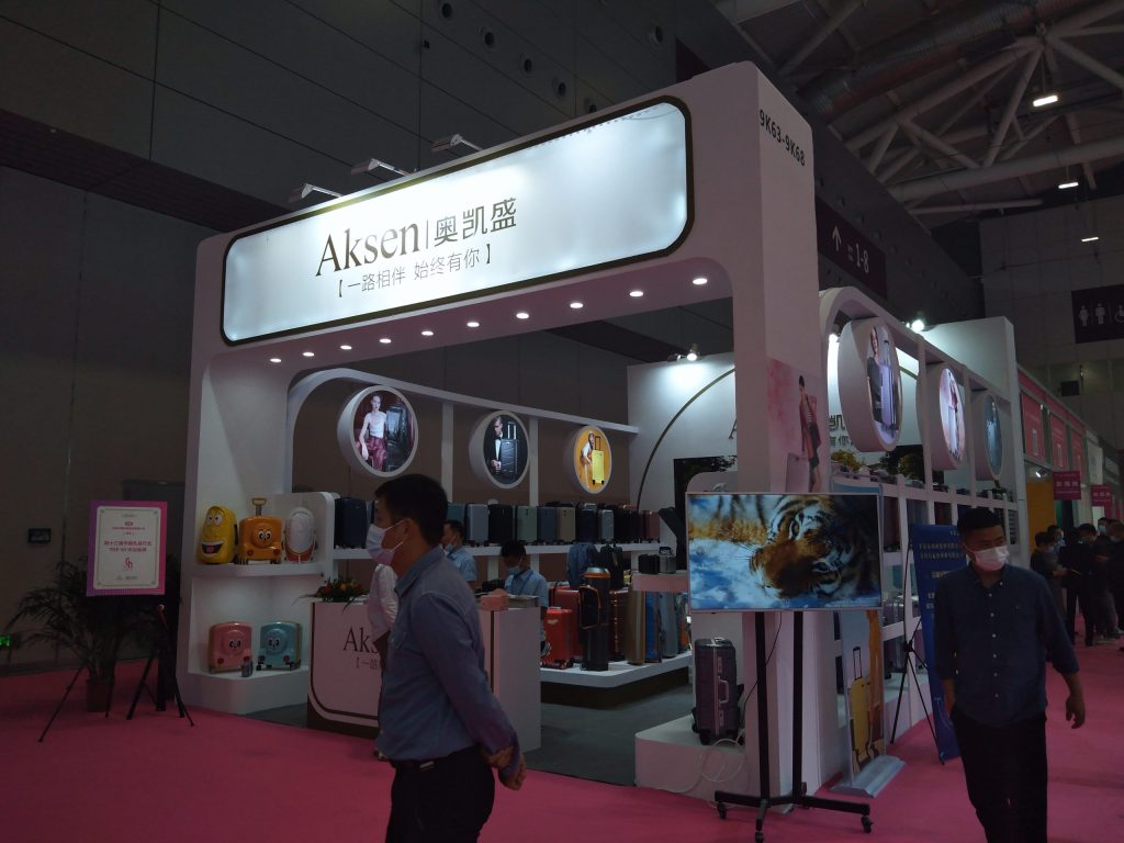 Busy trade show booth. People walk past displays of various luggage, a large sign, and a monitor showing a tiger.