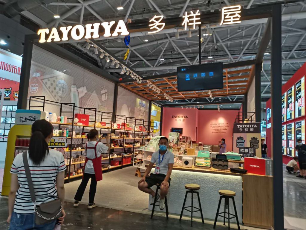 Exhibition booth showcasing many products on shelves. A masked man sits on a stool at a counter, while another person stands to the left.