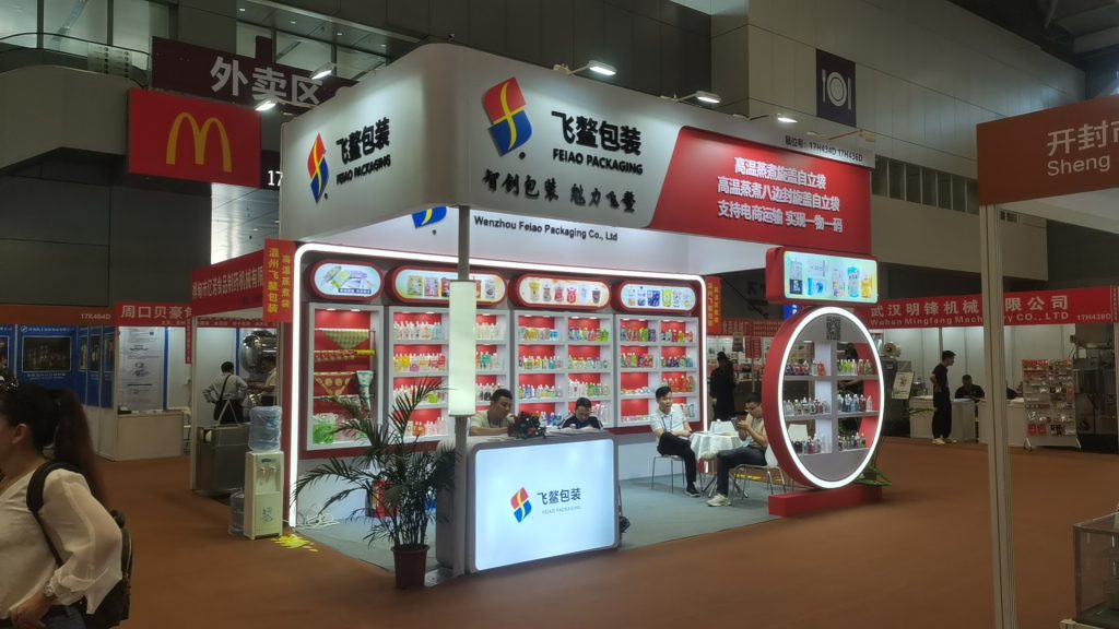 A brightly lit trade show booth with red and white accents, showcasing packaged products on shelves, with staff and visitors inside.