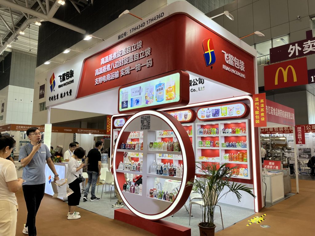 Exhibition hall view of a red and white trade show booth displaying packaged goods, surrounded by walking attendees.