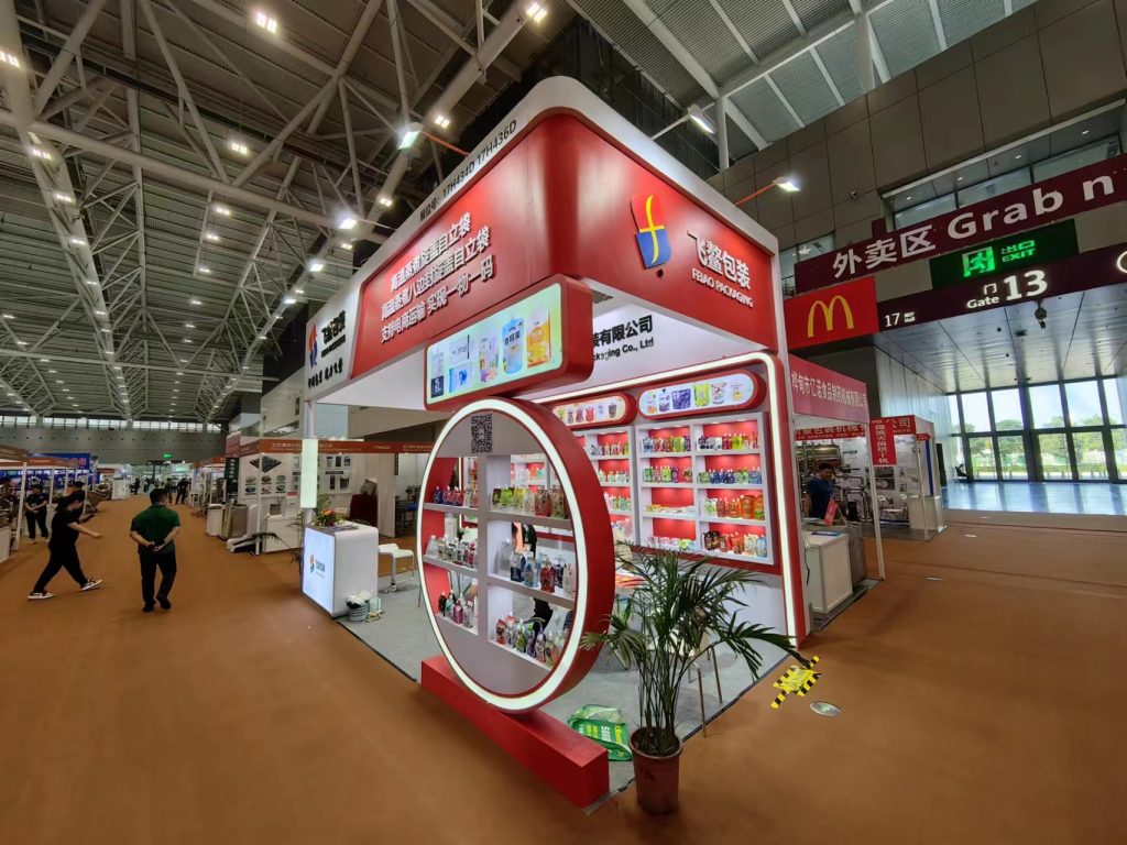 Wide shot of a red and white trade show booth filled with products, surrounded by attendees in a large exhibition hall.