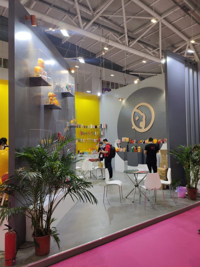 A modern exhibition booth with yellow and grey walls displays merchandise including yellow duck figures. Several people browse products near tables and chairs.
