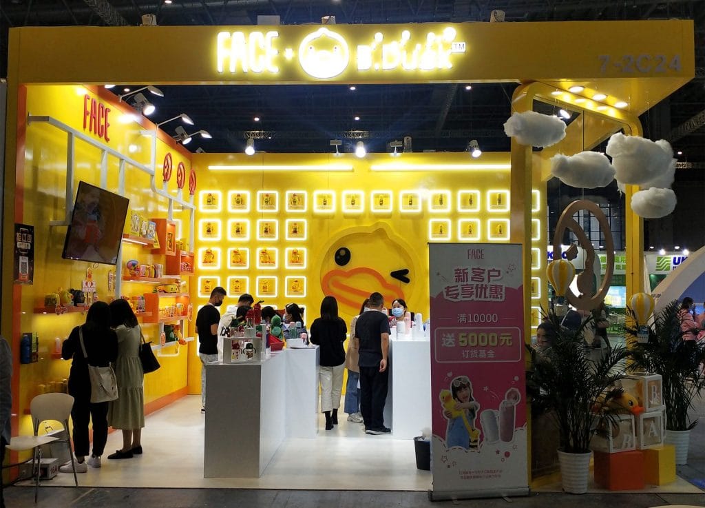Yellow exhibition booth. People browse products on shelves and counters, a large duck logo on the back wall.