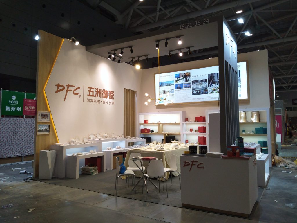 Modern exhibition booth with white display cases holding ceramic products and red boxes, a central table, screen, and contemporary lighting.