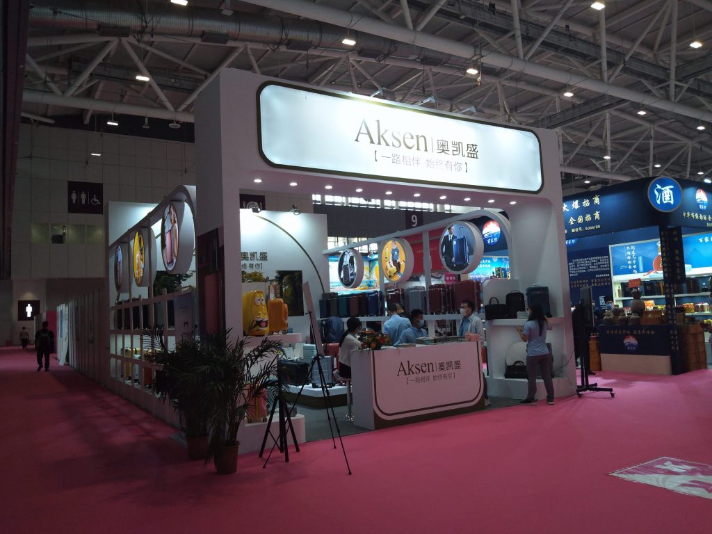 Wide view of a trade show booth with numerous luggage displays, staff at desks, and other exhibits in the background.
