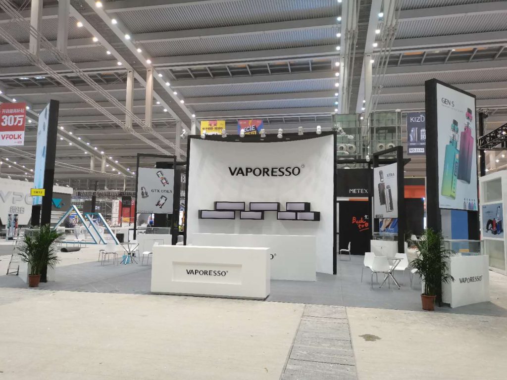A white exhibition booth with a large sign, display shelves, and a reception desk in a brightly lit convention hall.