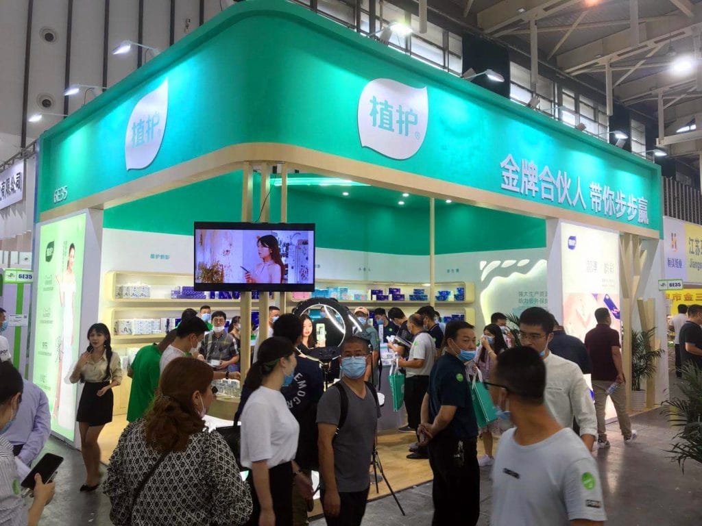 An indoor exhibition booth, crowded with masked attendees. The teal and white booth features a screen showing a woman using a phone.