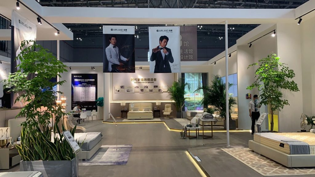 A spacious exhibition booth features beds, chairs, and potted plants, with two large banners of a man in a suit hanging above.