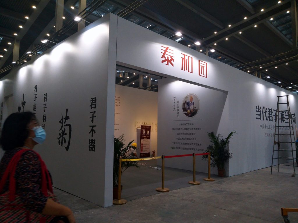 Woman in blue mask walks past a white exhibition booth with Chinese characters, a portrait, and an arched entrance.