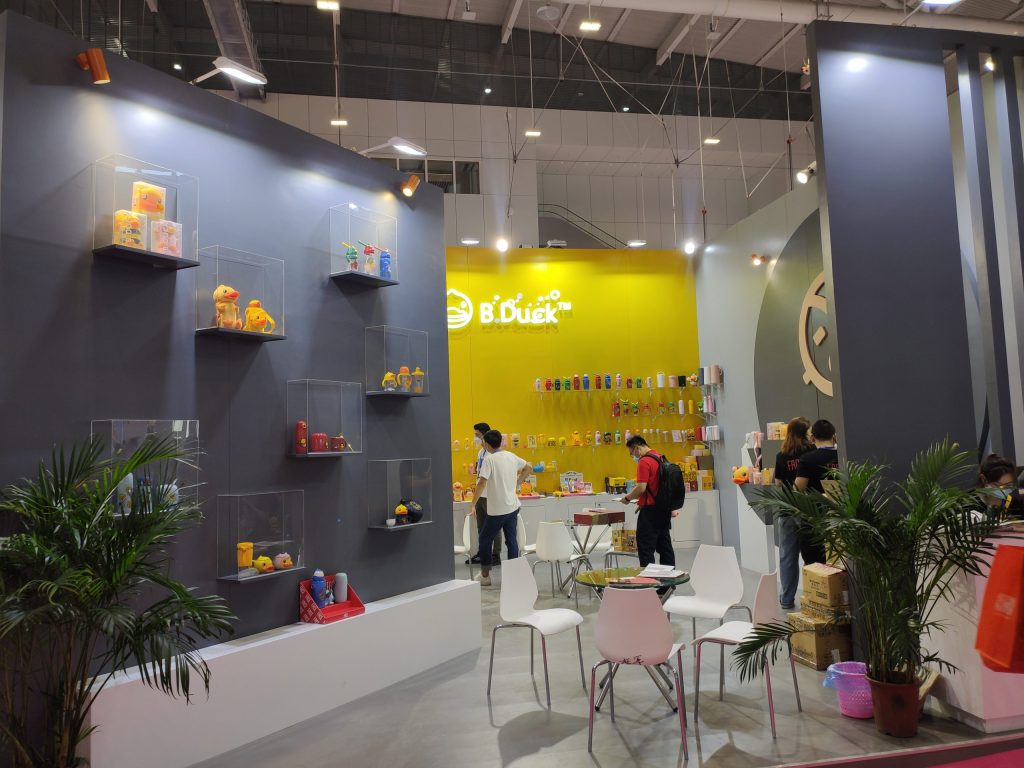A vibrant trade show booth features yellow and grey walls with shelves of products. Attendees observe merchandise and interact near small tables.
