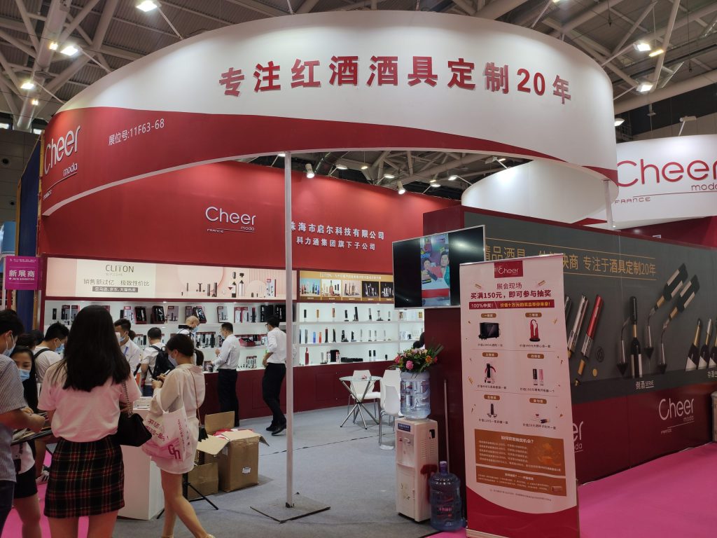 People browse a red and white exhibition booth displaying numerous wine accessories. A large overhead banner is visible.