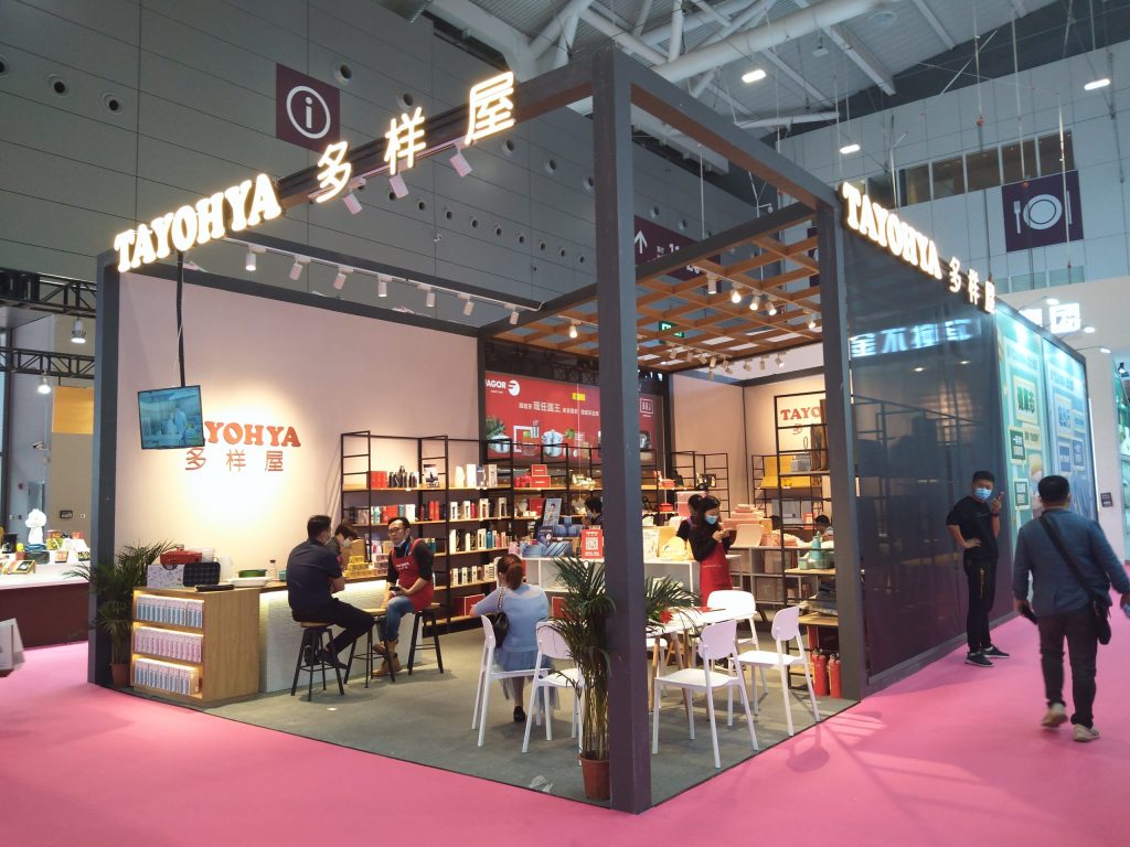 Live photo of a bustling exhibition booth with products on shelves, a counter, white tables, and chairs. Several visitors are browsing and interacting. Pink carpet is on the floor.