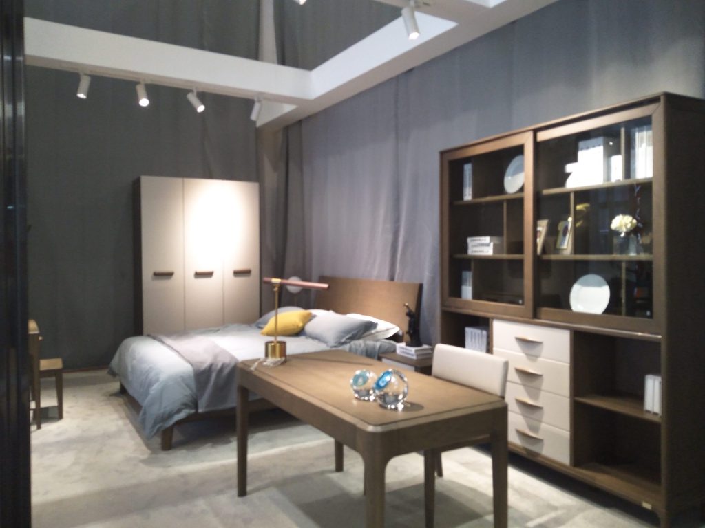A bedroom display featuring a bed with grey bedding, a wooden desk with two glass globes, and a tall storage cabinet.