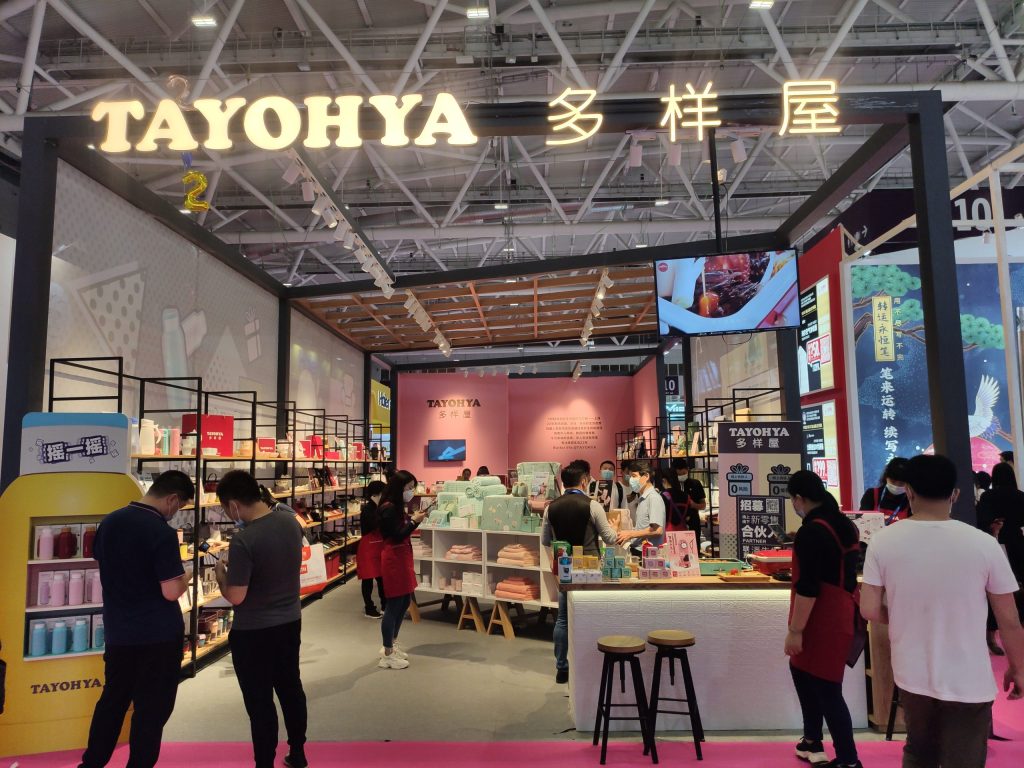 Busy exhibition booth with a pink floor, filled with masked visitors and staff in red aprons. People browse various displayed products.