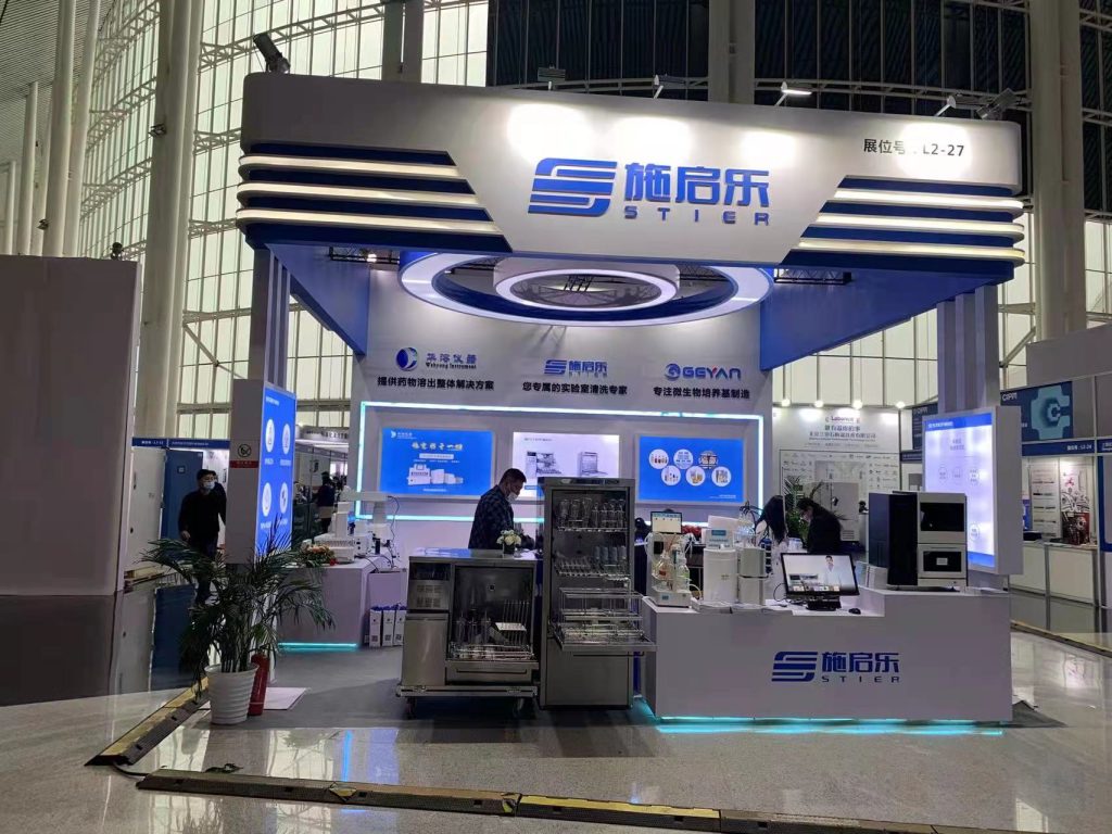 Real-world exhibition booth with blue and white accents, screens, products, and people in a brightly lit convention hall.