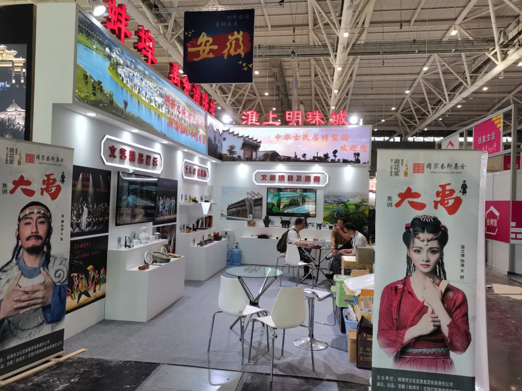 Busy exhibition booth with large screens showing landscapes and cultural scenes. Several people sit at a table, and banners feature traditional figures.